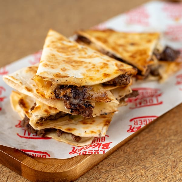 A stack of cheesy beef quesadillas on Local Bigger Burger branded paper, on a wooden board.