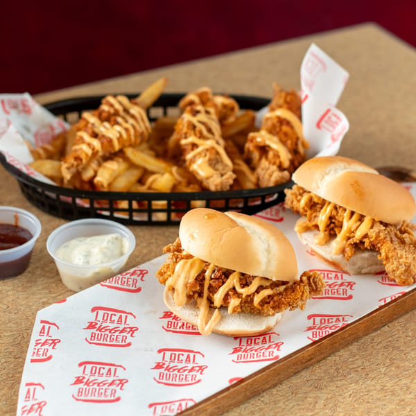 Local Bigger Burger's fried chicken sandwiches with sauce, a basket of fries, and dipping sauces.