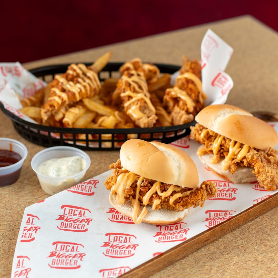 Local Bigger Burger's fried chicken sandwiches with sauce, a basket of fries, and dipping sauces.