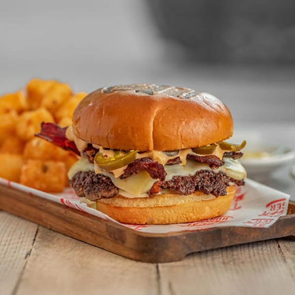 A Local Bigger Burger with bacon, jalapeños, and cheese, served with tater tots on a wooden tray.