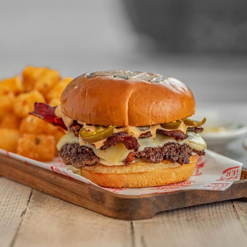 A Local Bigger Burger with bacon, jalapeños, and cheese, served with tater tots on a wooden tray.