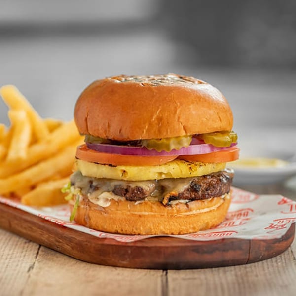 A Bigger Burger with pineapple, red onion, tomato, pickles, and fries on a wooden board.
