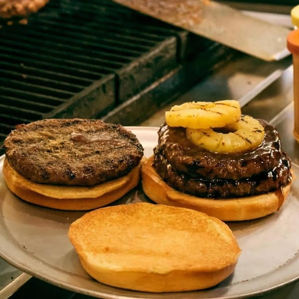 Two Bigger Burgers, one with pineapple, and a bun on a metal tray near a grill.